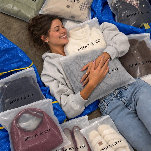 Smiling woman lying among Bisous & Co packages: coats, knits, a burgundy woven bag, ballet flats and cozy slippers