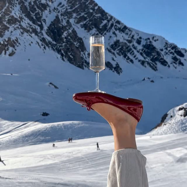 Capri Flat in burgundy held up over a snowy ski slope, with a glass of champagne balanced on top