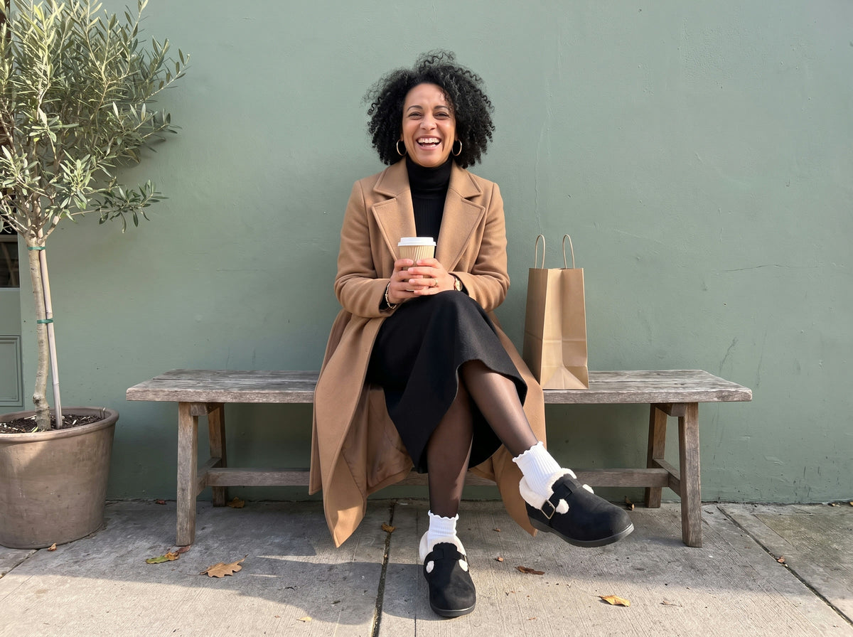 A woman laughing on a bench with a coffee, black faux-fur clogs over ruffle socks on a dry sidewalk