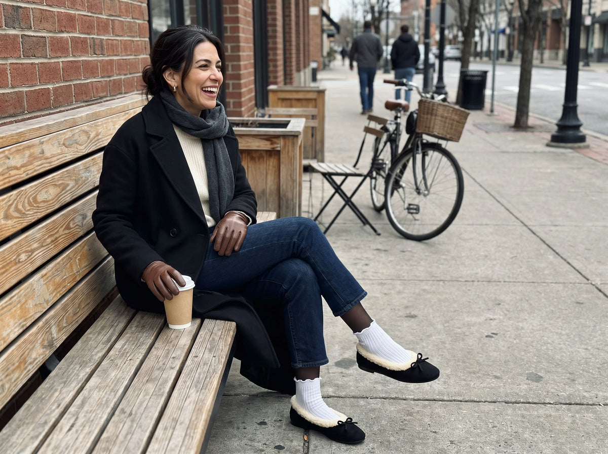 A woman laughs on a city bench in winter wearing the Courchevel Flat in Black with white ruffle socks over sheer black tights