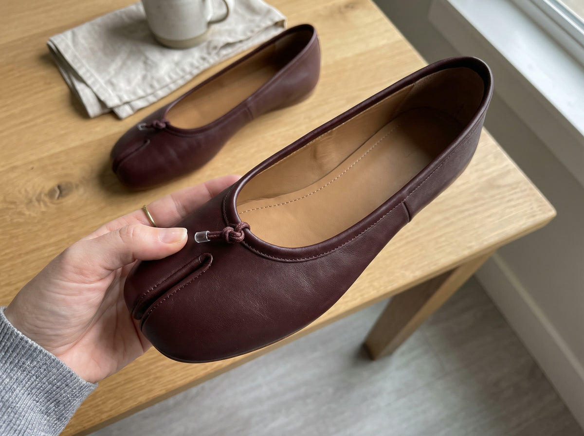 A hand holds an Oxblood split-toe flat to show the bound and stitched split and the caramel lining