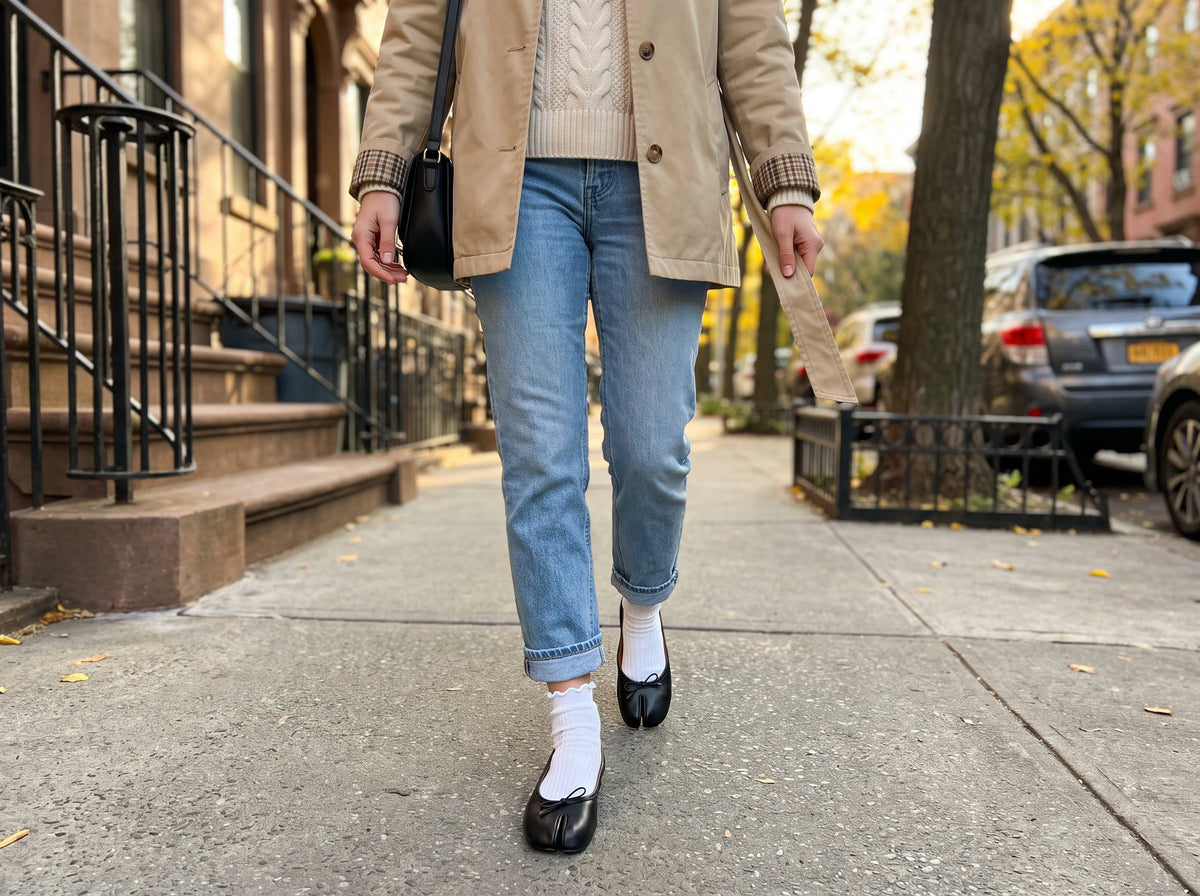 A short beige jacket, a cream knit, rolled jeans, white ruffle socks and black split-toe ballet flats on a city sidewalk