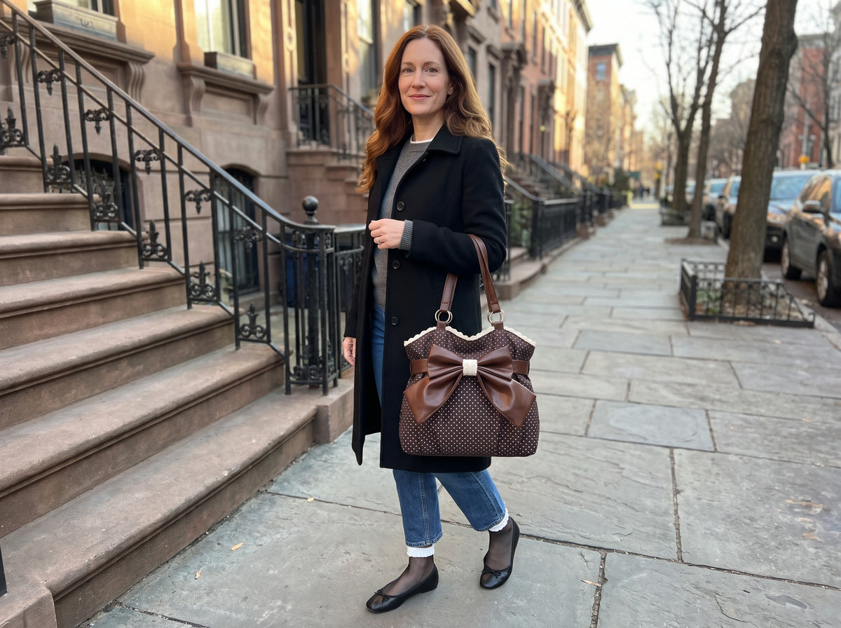 A woman in a black coat and jeans carries the Paris Bow Tote in Chocolate on her shoulder on a brownstone street