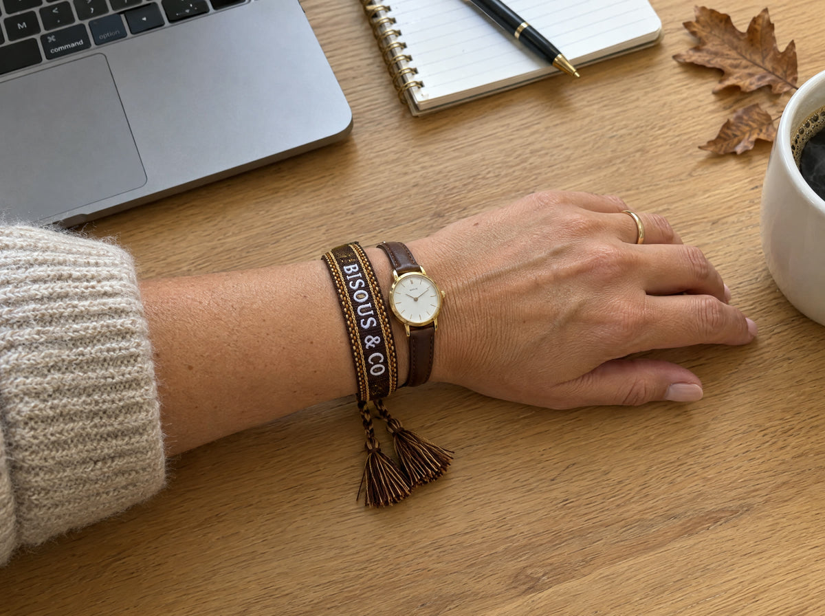 Woven bracelet in Chocolate Brown worn flat next to a slim gold watch, under the cuff of an oatmeal sweater, hand resting on a desk