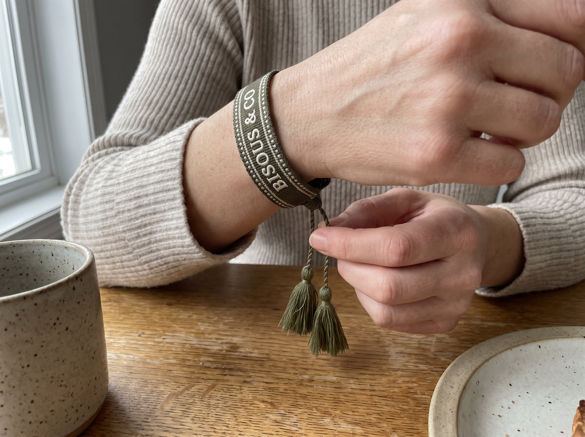A woman pulls the two tassels of her Olive Green woven bracelet to close the sliding knot, at the breakfast table on a fall morning